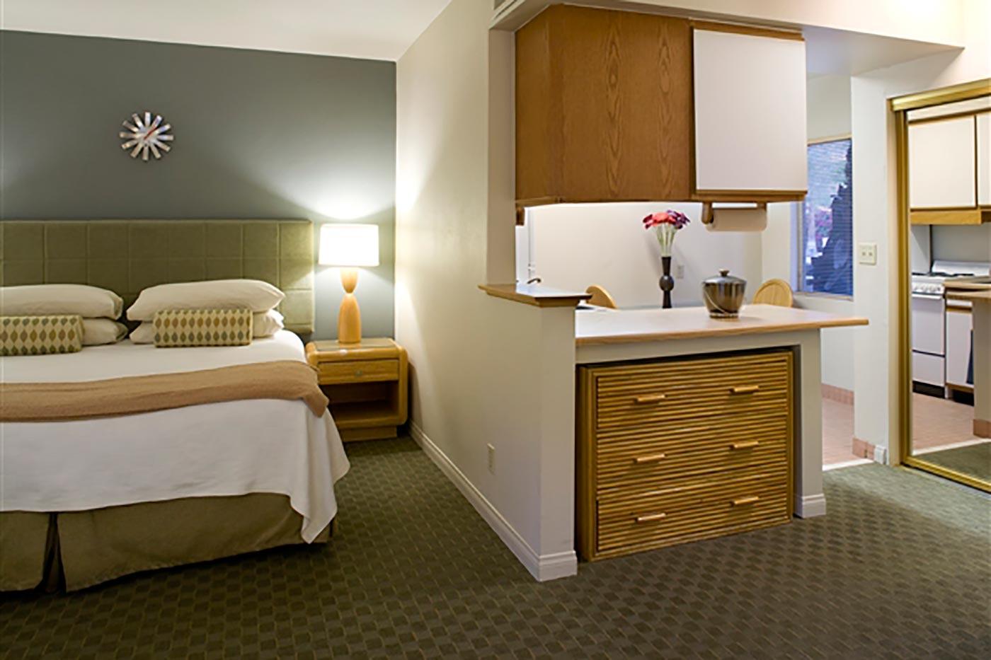 A hotel room with a bed, nightstand, lamp, and kitchenette.